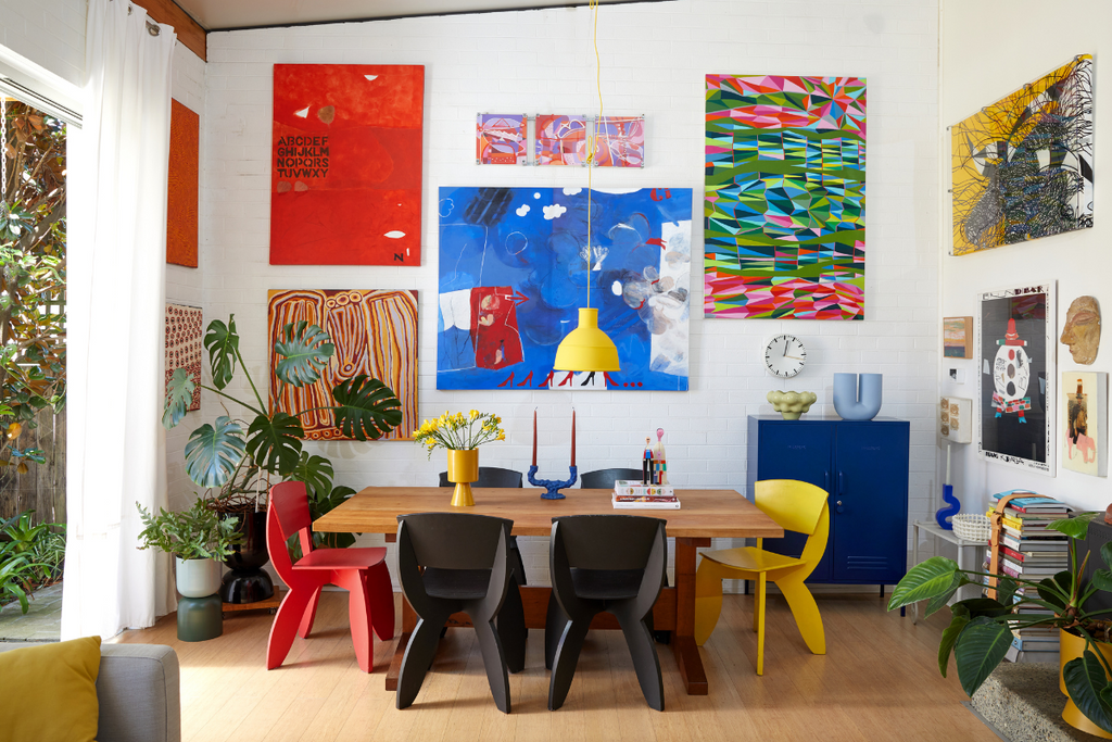 A colorful dining room featuring white brick walls, large abstract paintings and quirky chairs in red, black and yellow.