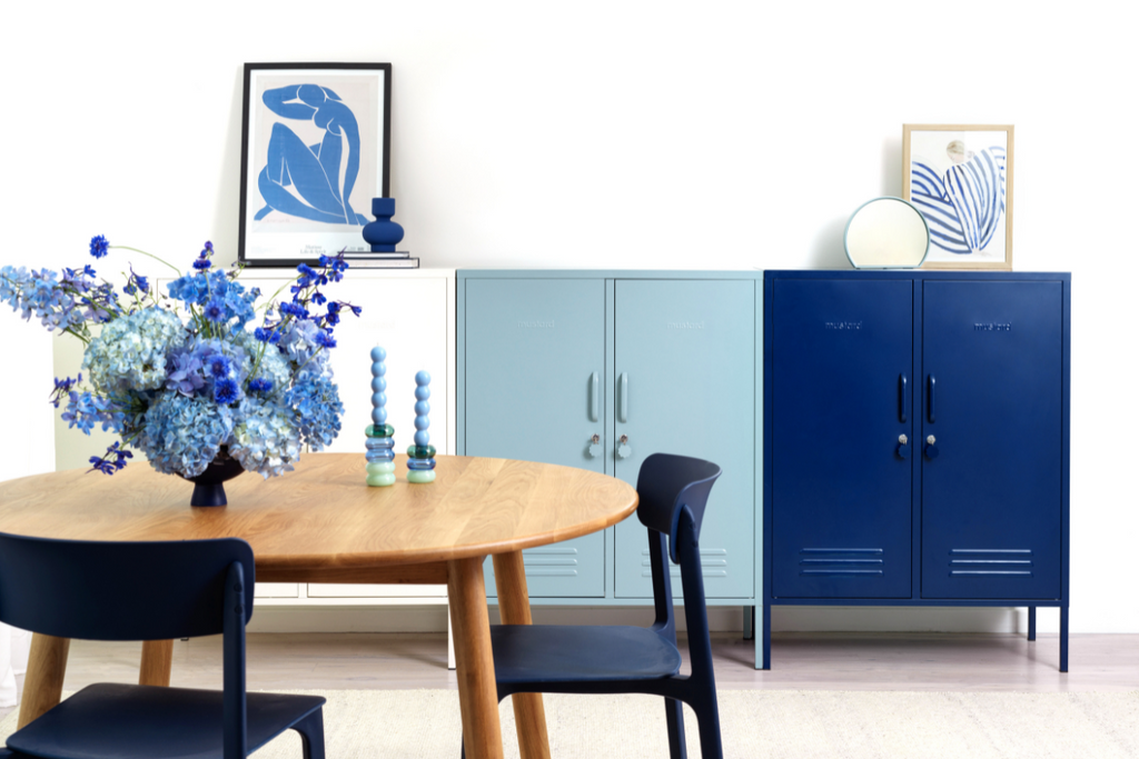 A trio of Midi lockers in an ombre pattern from white to Ocean to Navy are styled behind a round wooden dining table. There is a vase of luscious, full blue flowers and a selection of blue-toned artworks.