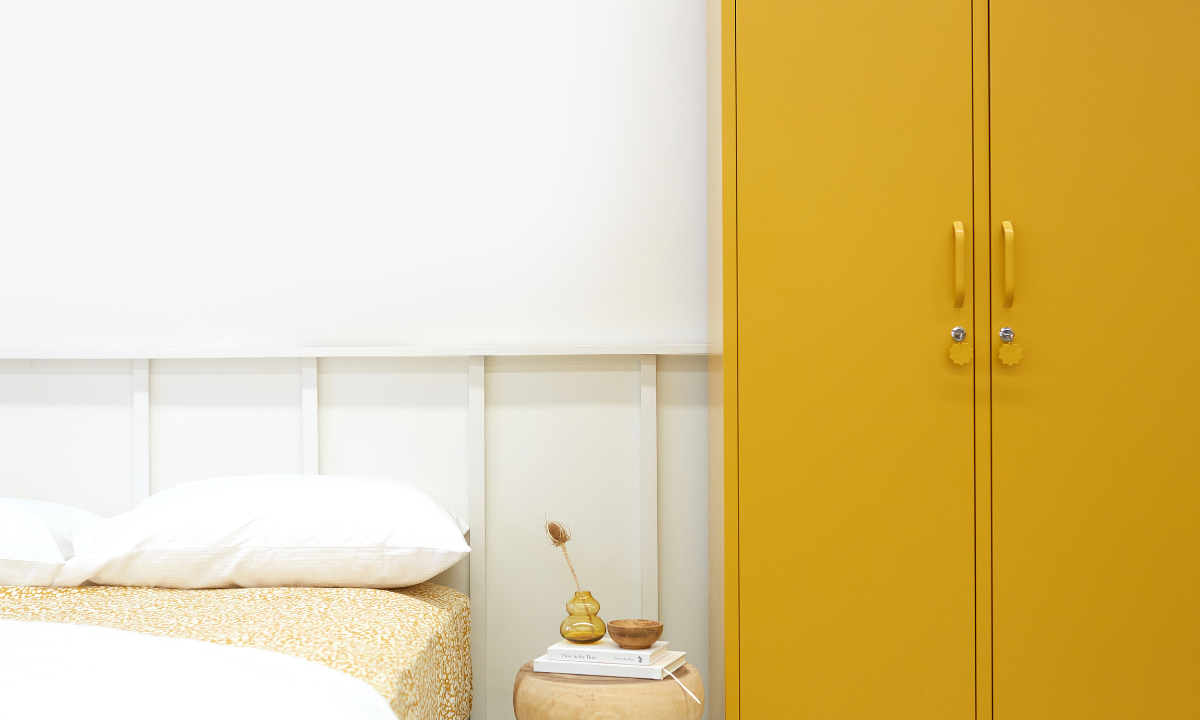 A Mustard yellow Twinny locker sits in a white room, next to a bed styled in neutral linens with a small wooden stool as a bedside table.