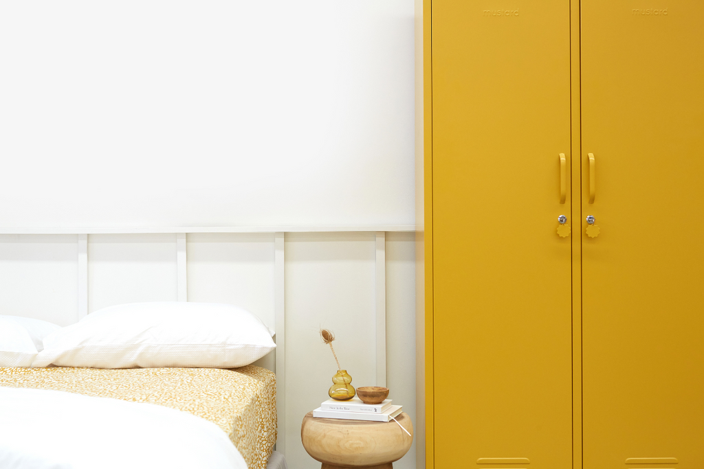 A Mustard yellow Twinny locker sits in a white room, next to a bed styled in neutral linens with a small wooden stool as a bedside table.