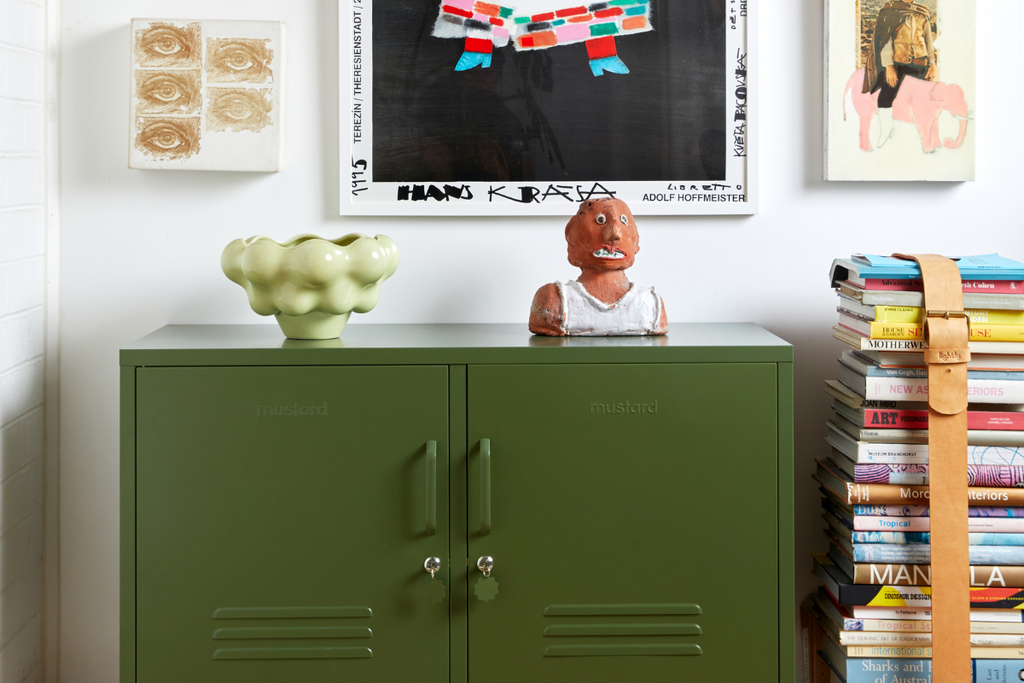An Olive Lowdown Mustard Made Locker is styled next to a stack of coffee table books and an eclectic range of colorful artworks.