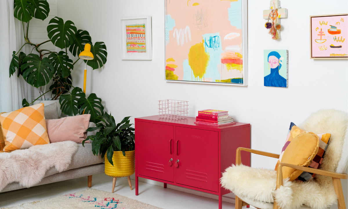 A Poppy red Lowdown sits in a white room styled with warm yellow and pink accents and art. There is a large Monstera plant in the corner.
