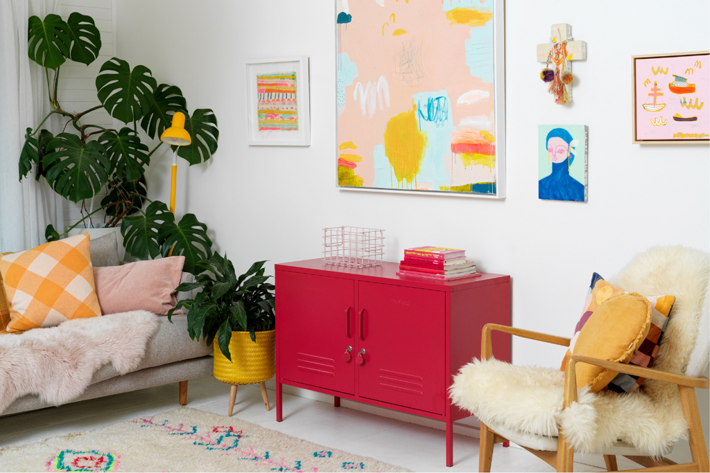 A Poppy red Lowdown sits in a white room styled with warm yellow and pink accents and art. There is a large Monstera plant in the corner.